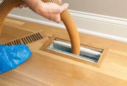 A worker is using a shop vacuum to clean a vent and duct on a new hardwood floor. The blue plastic is a protective cover over the worker's shoe.