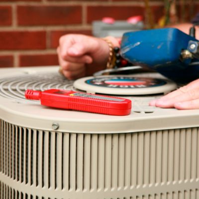 Close up of an AC condenser unit with tools and a man's hands resting on top